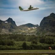 A small airplane flying over lush forest and rugged mountains under a cloudy sky.