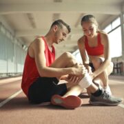 Side view of athlete sitting on rubber track in sports hall while sportive female partner helping to band leg after injury