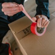 Focus on hands sealing a cardboard box with red tape, depicting careful packaging.